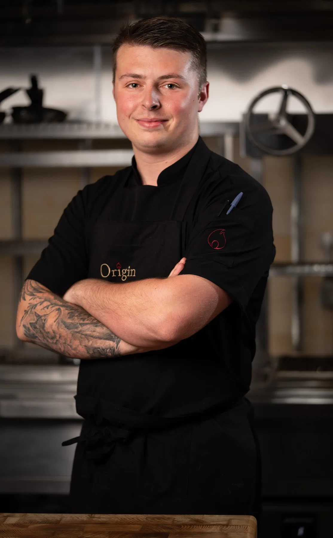 Chef in dark uniform, hands crossed, working kitchen behind